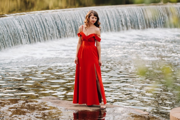 Ragazza in un lungo abito rosso vicino al lago nel parco al tramonto.