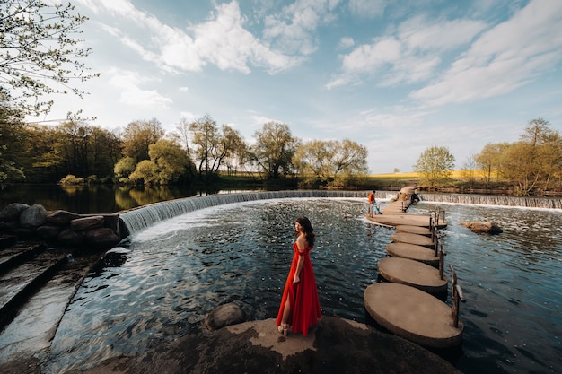 Ragazza in un lungo abito rosso vicino al lago nel parco al tramonto.