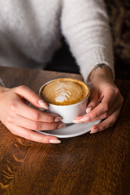 Ragazza in un maglione caldo bianco si siede al tavolo con in mano una tazza di cappuccino caldo con pista tra le mani. Bevanda al caffè