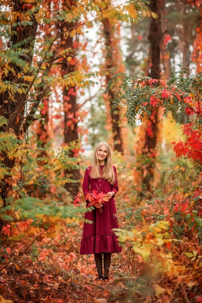 Ragazza in un vestito rosso nella foresta autunnale. Un bellissimo bosco da favola. Un mazzo di foglie nelle tue mani