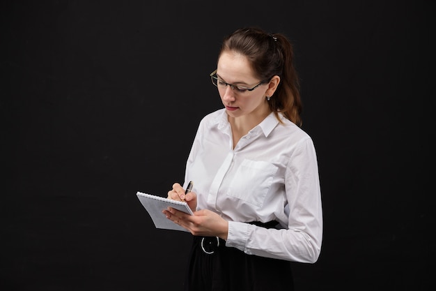 Ragazza in una camicia bianca con gli occhiali fa un'iscrizione in un notebook su uno sfondo nero.