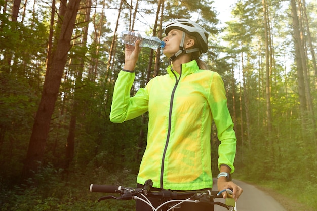 Ragazza in una giacca a vento verde brillante, pantaloncini, casco e occhiali che beve acqua dopo l'allenamento