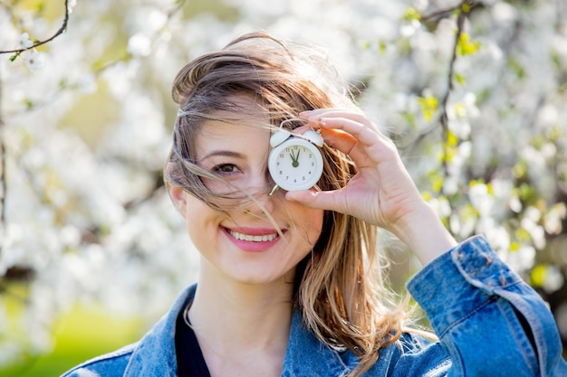 Ragazza in una giacca di jeans e sveglia si trova vicino a un albero in fiore nel parco. Stagione primaverile