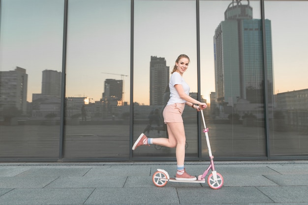 Ragazza in una maglietta bianca cavalca uno scooter sullo sfondo di un muro scuro, guarda la telecamera e sorride.