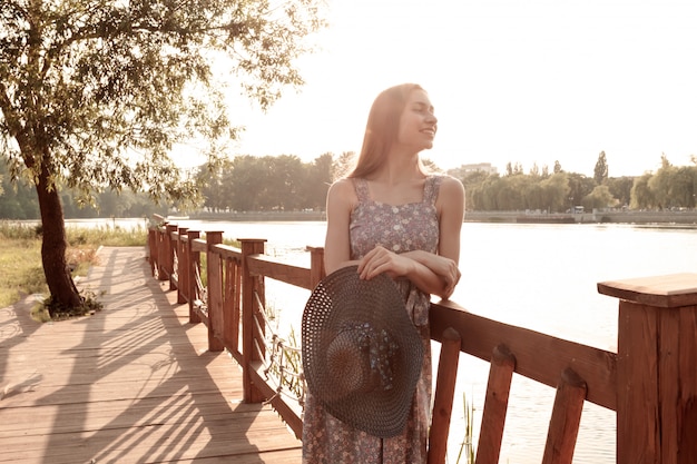 Ragazza in vestito con capelli lunghi che posano vicino all'acqua. Ritratto di estate della ragazza al tramonto.