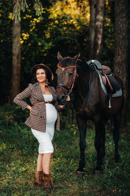 Ragazza incinta con una grande pancia in un cappello accanto ai cavalli nella foresta in natura. Elegante ragazza in abiti bianchi e una giacca marrone.