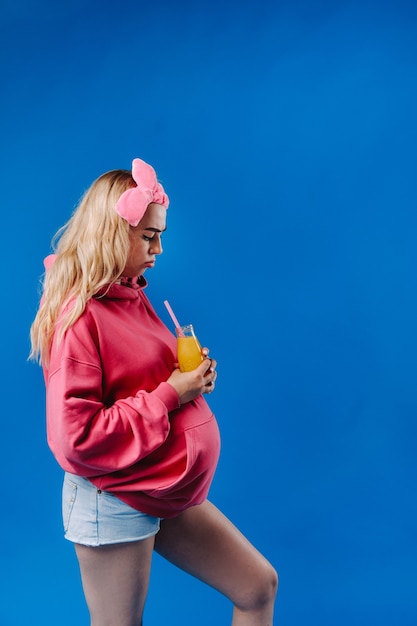 Ragazza incinta in abiti rosa con una bottiglia di succo su sfondo blu.