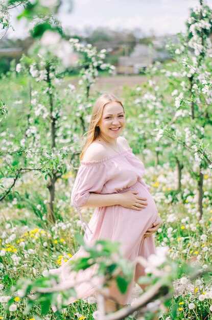 Ragazza incinta in un vestito rosa in un giardino fiorito con alberi e fiori verdi