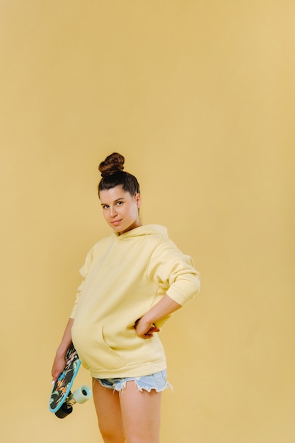 Ragazza incinta in una giacca gialla con uno skateboard in mano su uno sfondo giallo