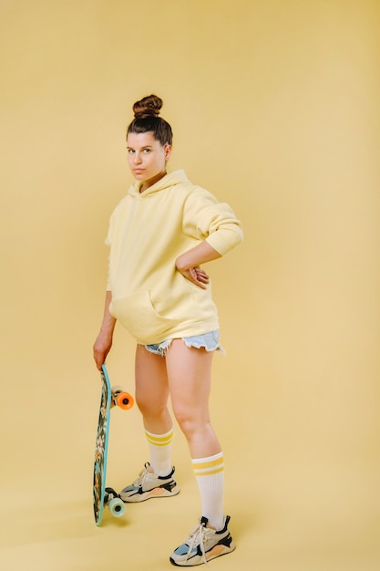 Ragazza incinta in una giacca gialla con uno skateboard in mano su uno sfondo giallo