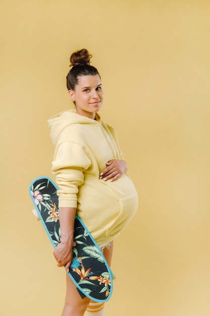 Ragazza incinta in una giacca gialla con uno skateboard in mano su uno sfondo giallo.