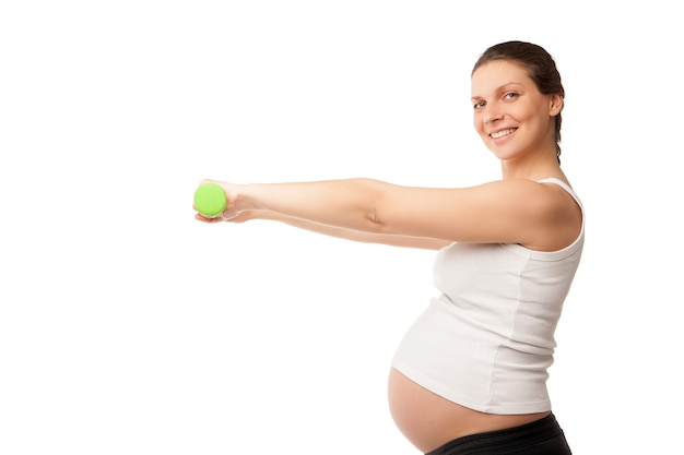 Ragazza incinta sportiva che solleva manubri isolati su sfondo bianco, allenamento al chiuso, sport per donne in attesa, concetto di gravidanza sana