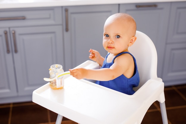 Ragazza infantile che si siede nella sedia alta del bambino che mangia su una priorità bassa bianca. Concetto di cibo per bambini, spazio per il testo