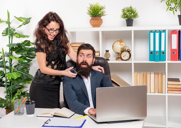 Ragazza ingannevole spaventa l'uomo barbuto che taglia la barba con le forbici sul posto di lavoro in ufficio, barbiere.