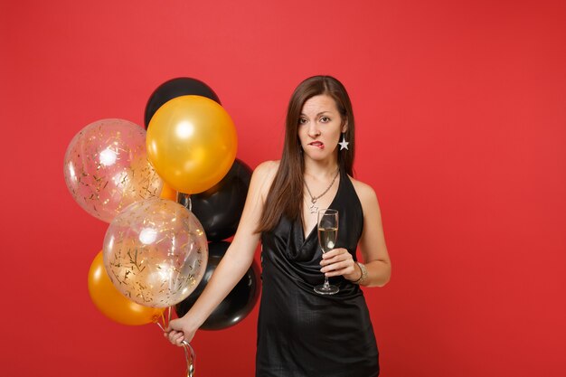 Ragazza insoddisfatta in abito nero che celebra mordendosi le labbra tenendo in mano un bicchiere di mongolfiere di champagne isolate su sfondo rosso. Festa della donna Felice Anno Nuovo compleanno mockup festa concetto.