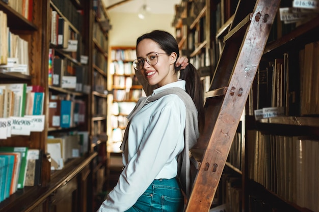 Ragazza intelligente che indossa una camicia bianca in piedi vicino agli scaffali della biblioteca