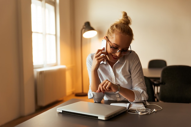 Ragazza intelligente con gli occhiali parlando al telefono e guardando il suo nuovo orologio