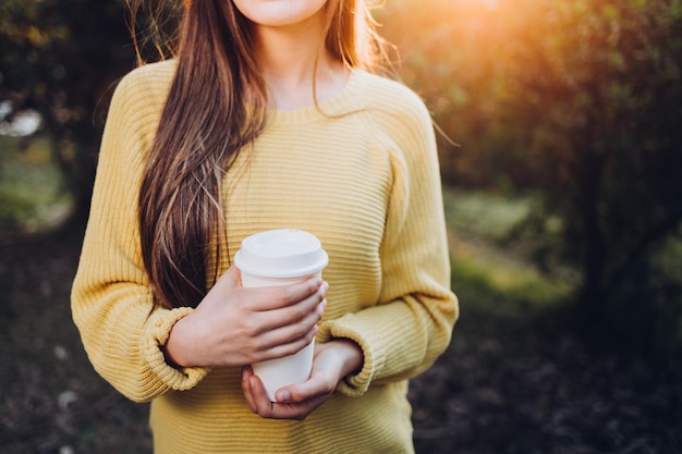 Ragazza irriconoscibile che indossa un maglione e tiene una tazza di caffè bianca nel parco.