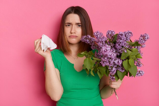 Ragazza malata con bouquet di fiori lilla soffre di malessere allergico e rinite malattia stagionale stanco di starnuti allergia alla fioritura tiene pose fazzoletto isolato su sfondo rosa parete