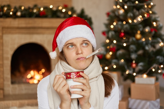 Ragazza malata in cappello della Santa che misura la temperatura mentre tiene una tazza di bevanda calda nelle mani, guardando da parte con espressione triste, sognando di festeggiare il nuovo anno, ubicazione vicino al camino e albero di Natale.