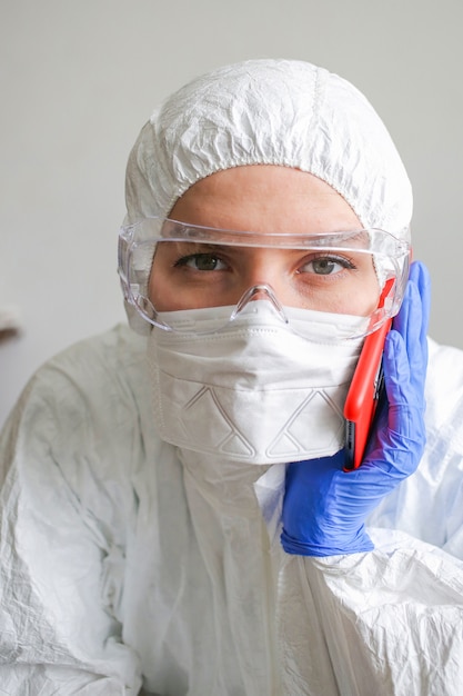 ragazza medico infermiera in una tuta protettiva e maschera e occhiali protettivi parlando al telefono