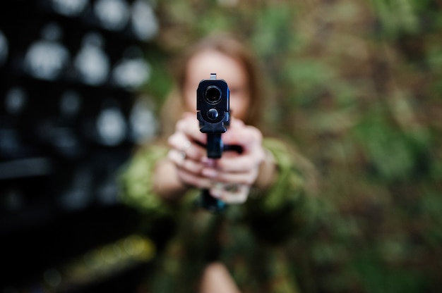 Ragazza militare in uniforme mimetica con la pistola a portata di mano contro il fondo dell'esercito su poligono di tiro. Concentrati sulla pistola.