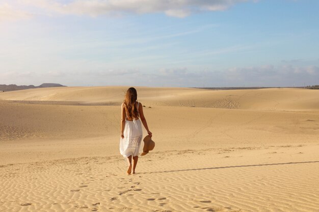 Ragazza misteriosa con vestito bianco e cappello che cammina sulla sabbia dorata delle dune del deserto al tramonto