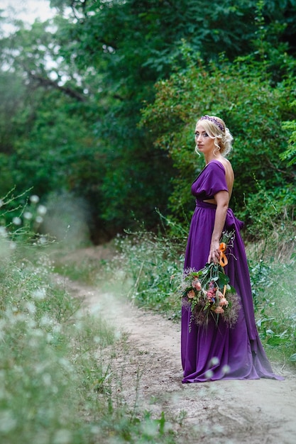 Ragazza modella bionda in un vestito lilla con un bouquet con una foresta verde