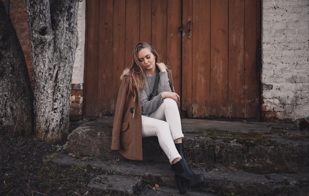 ragazza modello bionda in pantaloni bianchi, maglione lavorato a maglia grigio, stivali neri, cappotto marrone