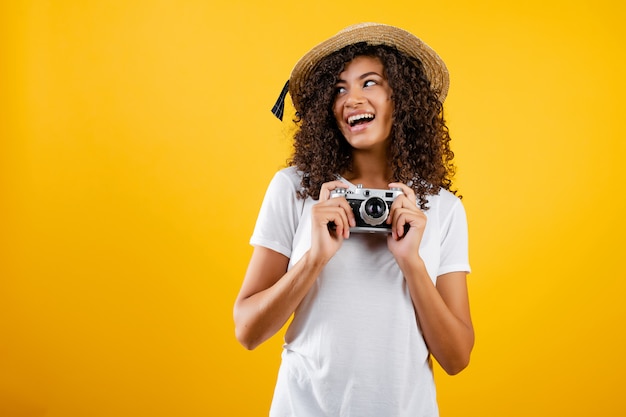 Ragazza nera alla moda dei pantaloni a vita bassa con la macchina fotografica d'annata e cappello isolato sopra giallo