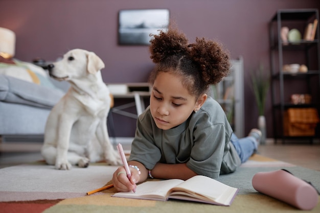 Ragazza nera che scrive nel diario sdraiato sul pavimento a casa con il cane sullo sfondo