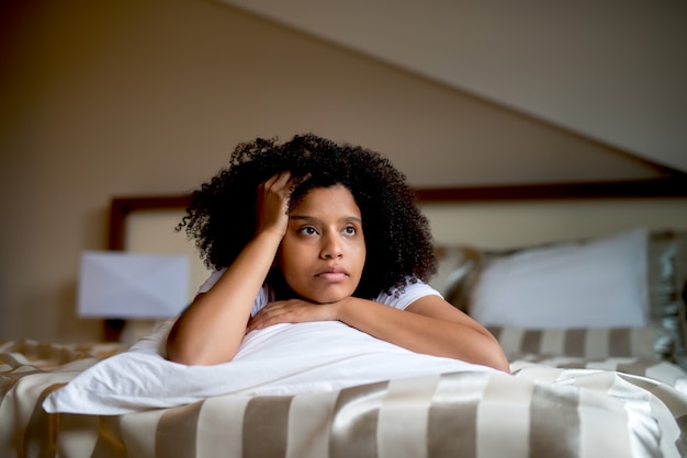 Ragazza nera riccia triste che si trova sul letto che abbraccia cuscino bianco.