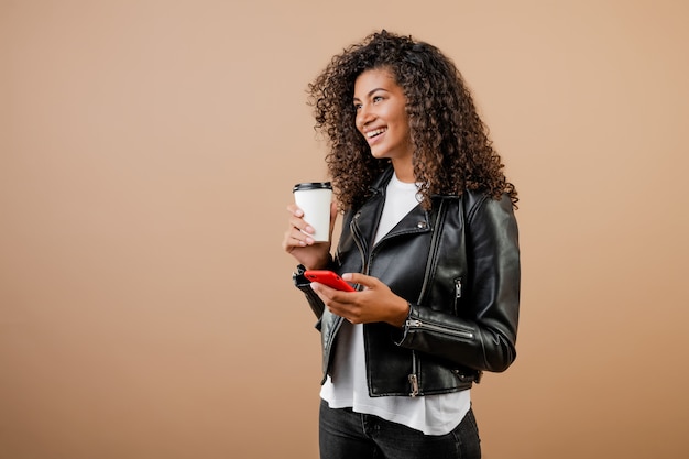 Ragazza nera sorridente felice con il telefono e il caffè per andare tazza isolata sopra marrone