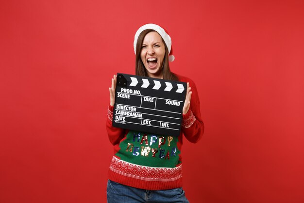 Ragazza pazza di Santa giovane con cappello di Natale che urla, con in mano un classico film nero che fa ciak isolato su sfondo rosso. Felice Anno Nuovo 2019 celebrazione festa concetto. Mock up copia spazio.