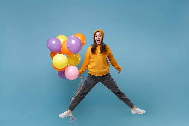 Ragazza pazza giovane donna in maglione e cappello in posa isolata su sfondo blu. Festa di compleanno, concetto di emozioni della gente. Mock up spazio di copia. Festeggiando tenendo palloncini d'aria colorati che saltano.