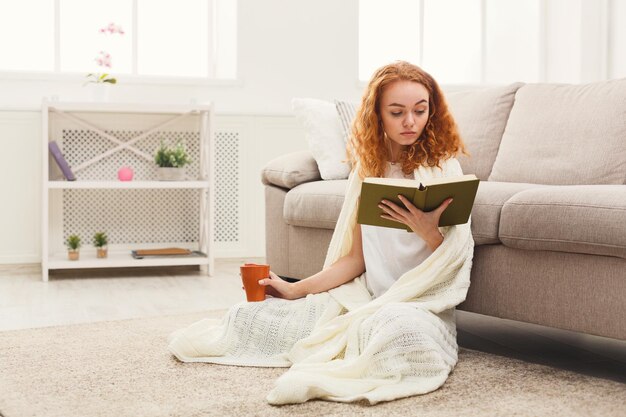 Ragazza pensierosa dello studente della testarossa che legge un libro. Giovane donna che studia e beve caffè a casa sul pavimento in una coperta bianca