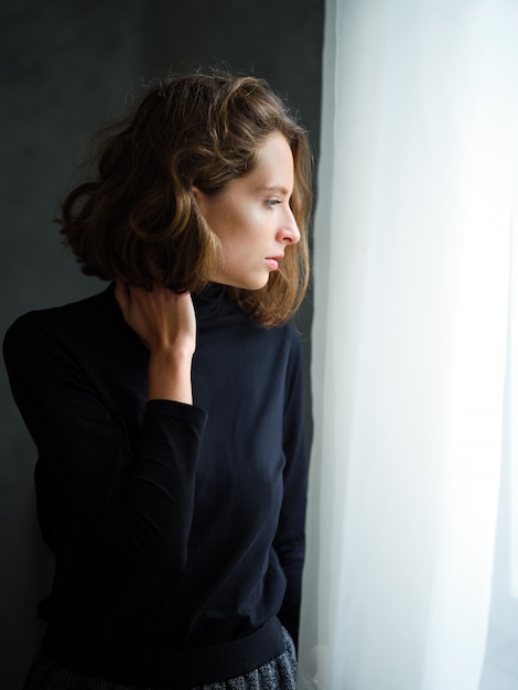 Ragazza pensierosa in pullover nero vicino alla finestra