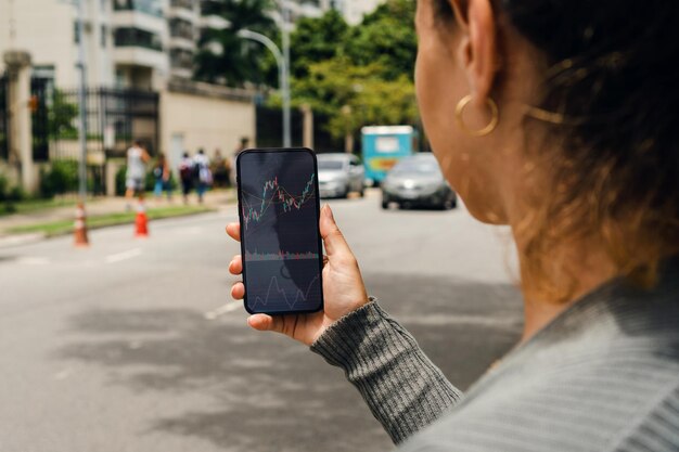 Ragazza per strada che tiene smartphone con il grafico del mercato azionario finanziario sullo schermo in attesa dell'arrivo dell'auto in borsa