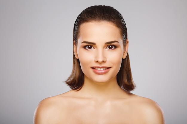 Ragazza perfetta con capelli castani, pelle fresca e pulita e spalle nude in posa al muro grigio, un modello con trucco nudo leggero, da vicino.