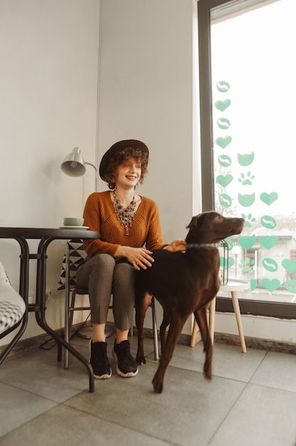 Ragazza positiva dei pantaloni a vita bassa che si siede al tavolo in un caffè luminoso, bere caffè e giocare con il cane