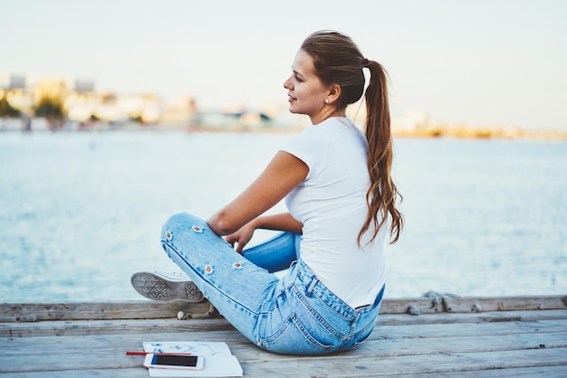 Ragazza positiva hipster in jeans alla moda che guarda da parte e riposa dallo studio sul lago giovane