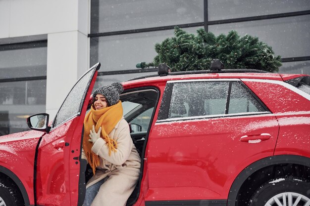 Ragazza positiva in piedi vicino all'auto con albero di Natale verde in cima.