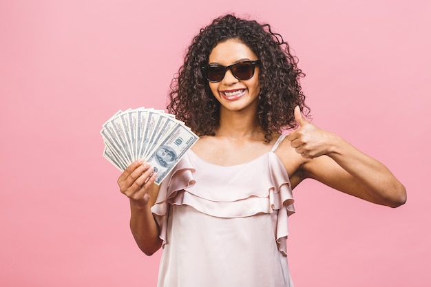 Ragazza ricca! Vincitore di soldi! Bella donna afroamericana sorpresa in vestito che tiene i soldi e guardando la telecamera isolata su sfondo rosa. Pollice su.