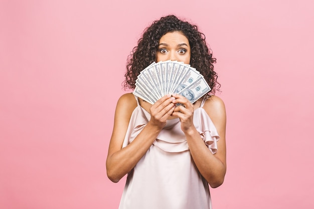 Ragazza ricca! Vincitore di soldi! Bella donna afroamericana sorpresa in vestito che tiene i soldi e guardando la telecamera isolata su sfondo rosa.