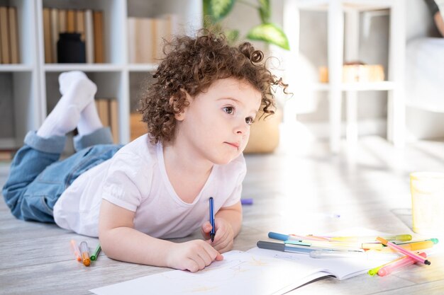 Ragazza riccia bambino disegno con pennarelli colorati sdraiato sul pavimento in soggiorno a casa