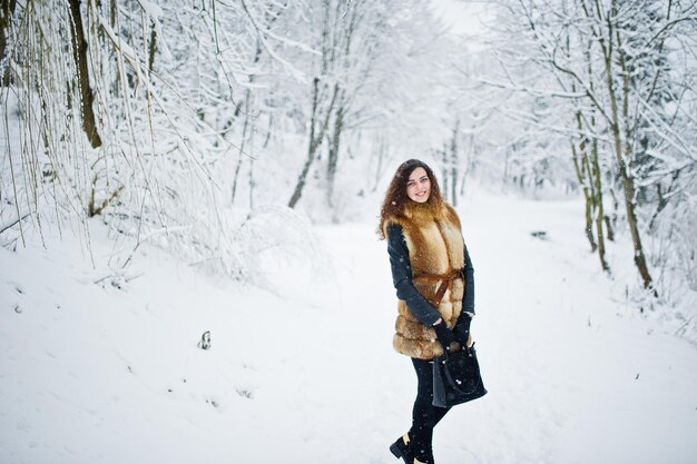 Ragazza riccia di eleganza in pelliccia e borsa al parco forestale innevato in inverno.