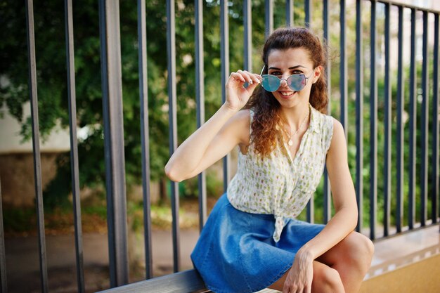 Ragazza riccia ed elegante indossa una camicetta con gonna di jeans blu seduta su una panchina Ritratto per le strade della città