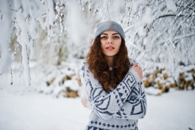 Ragazza riccia sveglia in maglione e cappelleria a Forest Park nevoso all'inverno.