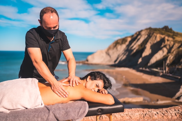 Ragazza rilassata nel massaggio sulla costa vicino al mare, massaggiatrice con maschera facciale nella pandemia di coronavirus