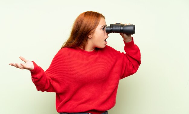 Ragazza rossa adolescente con maglione sopra verde isolato con binocolo nero